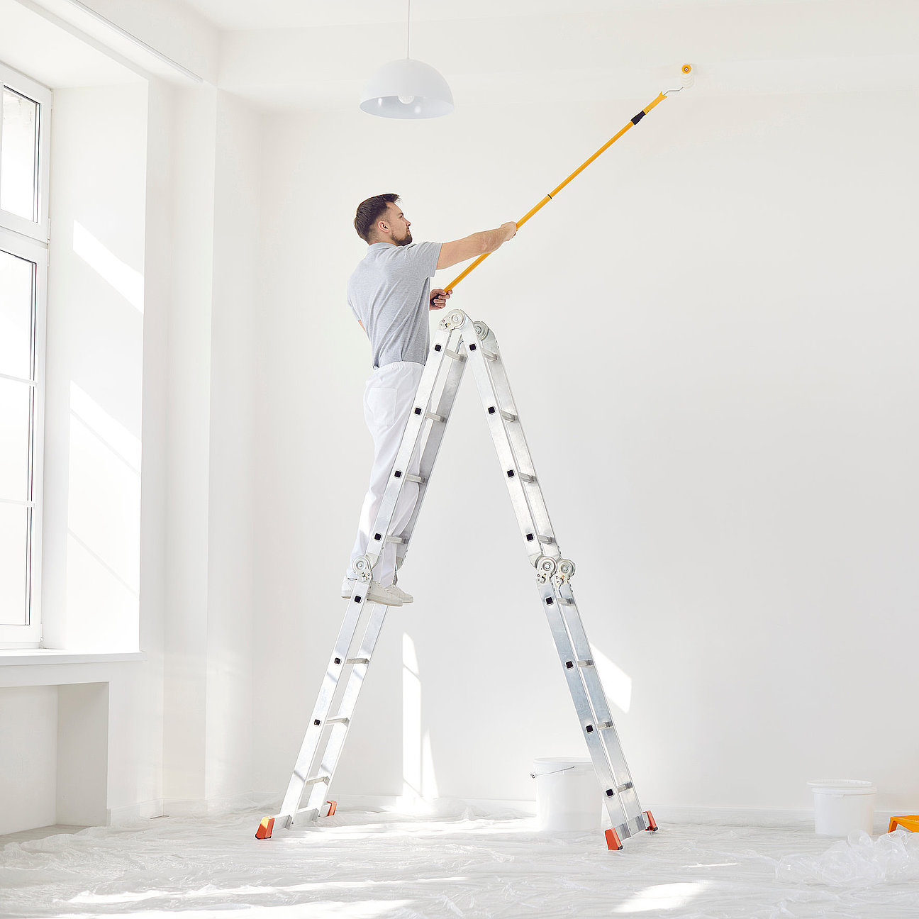 Portrait of young man painter from repair service in uniform standing on a ladder in empty room and painting the ceiling with paint roller. Renovation and renovating house concept. Bauarbeiter beim Spachteln einer Altbauwand - präzise Handarbeit