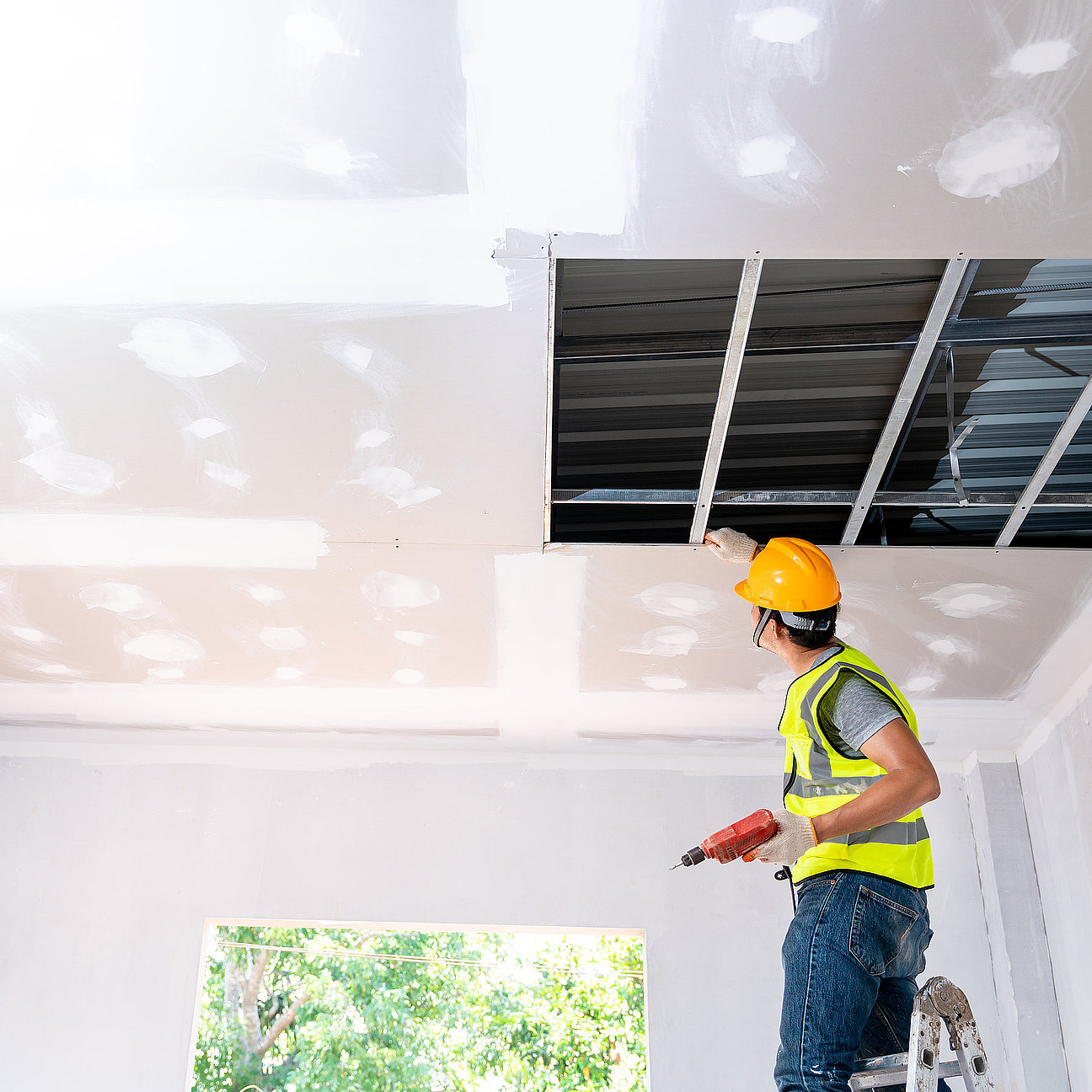 Asian construction worker assemble a suspended ceiling with drywall and fixing the drywall to the ceiling metal frame with screwdriver. Renovation, construction and ceiling installation concept. Bauarbeiter montiert abgehängte Decke